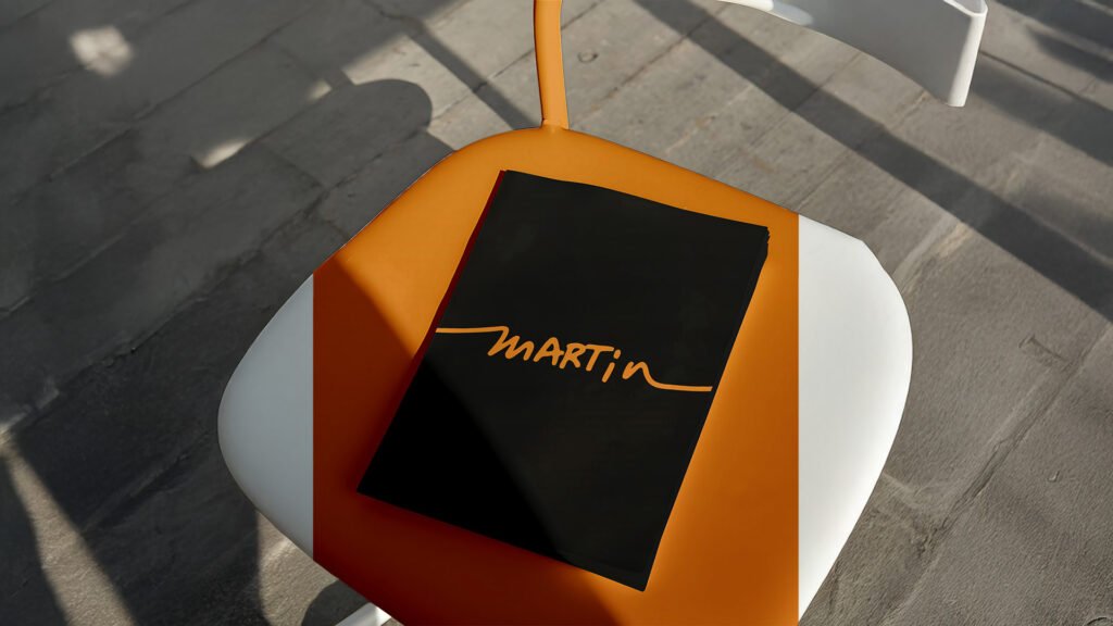 Martin-branded black folder with orange script logo placed on a half-painted orange and white chair in outdoor sunlight.
