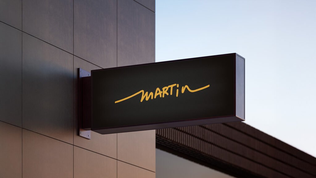 Illuminated black rectangular sign with the Martin logo in bold yellow script, mounted on a modern building facade at dusk.