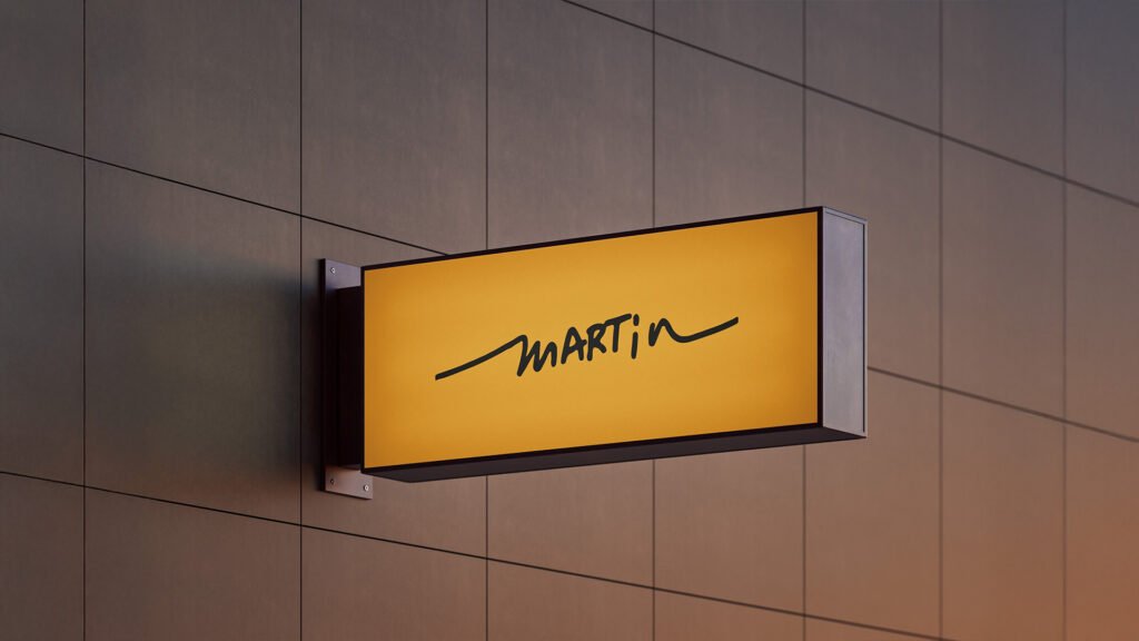 Illuminated yellow rectangular sign with the Martin logo in black handwritten script, mounted on a modern tiled building wall.