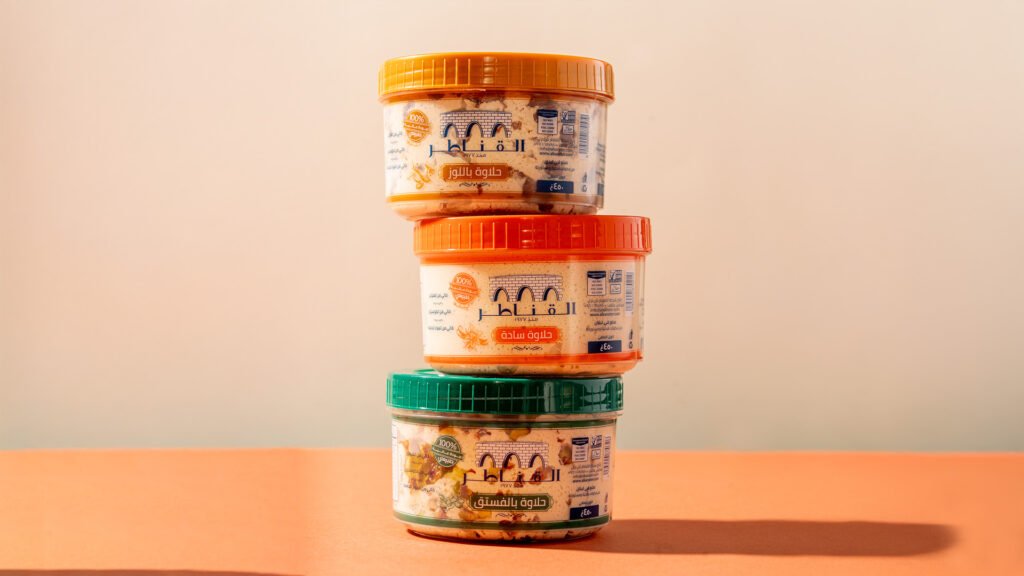 Stacked Al Kanater Halawa containers (plain, pistachio, almond) on a warm-toned background, showcasing traditional Middle Eastern sesame-based sweets for food branding and marketing.