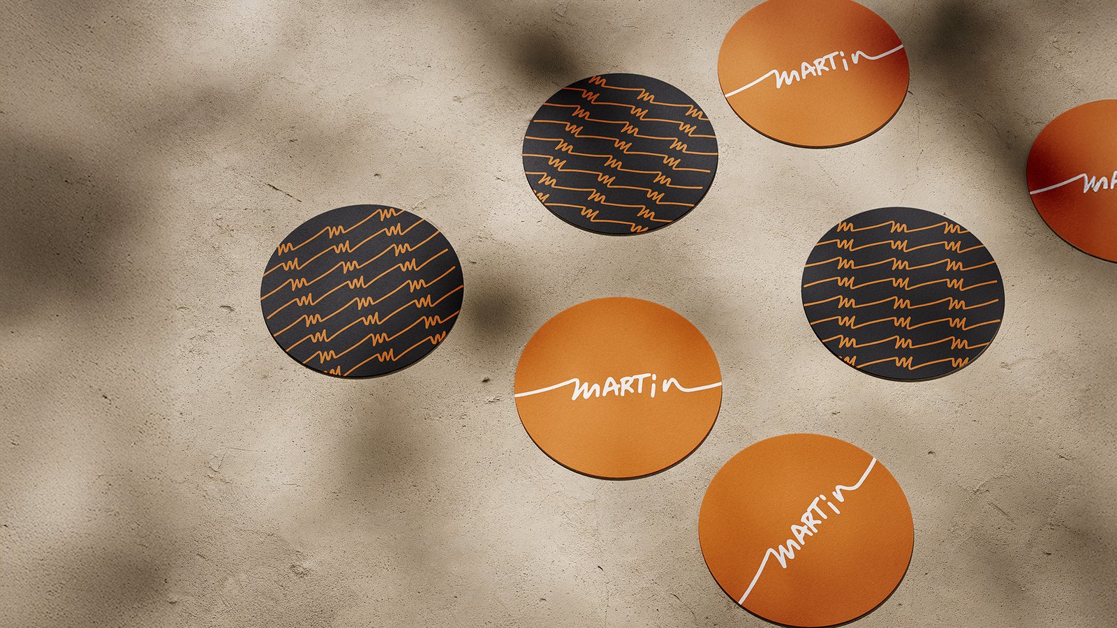 Branded circular coasters with the Martin logo and signature pattern in orange and black, displayed on a textured concrete surface with soft shadows.