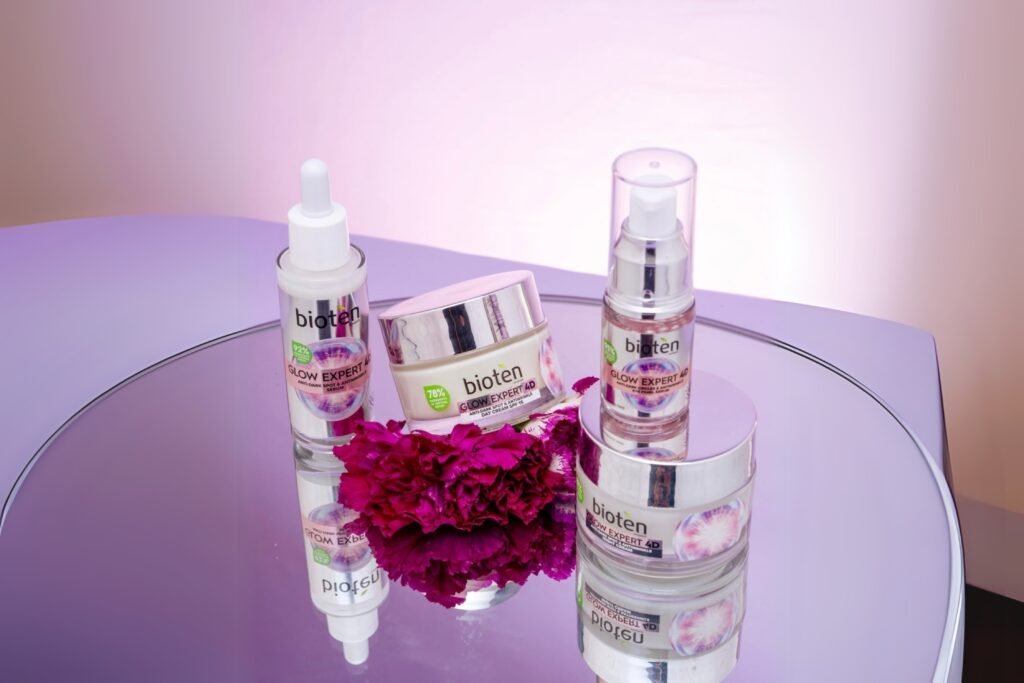 Bioten Glow Expert 4D skincare collection including serum, moisturizer, and day cream, displayed on a mirrored surface with floral accents for beauty product branding and campaign visuals.
