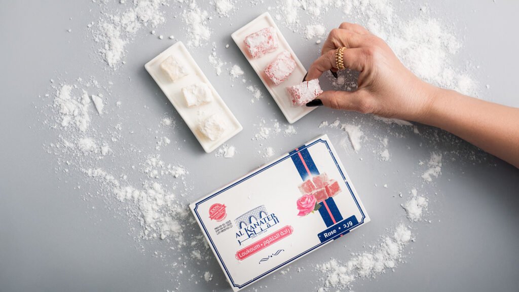 Hand picking a powdered sugar-covered rose loukoum (raha) from a white tray, with Al Kanater packaging and Turkish delight sweets styled on a gray background.